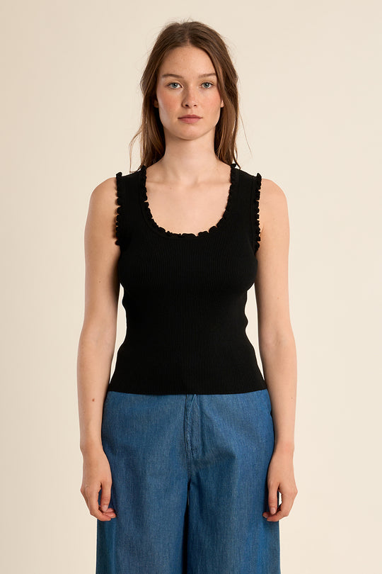 Woman wearing a black sleeveless top with ruffled details and blue pants on a beige background