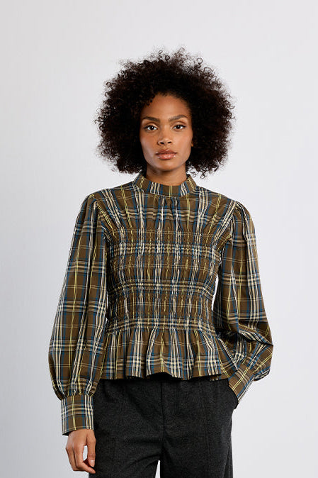 A model stands against a light gray studio background wearing a long-sleeve plaid blouse in olive green, yellow, and blue tones, paired with dark charcoal wide-leg trousers and black shoes.