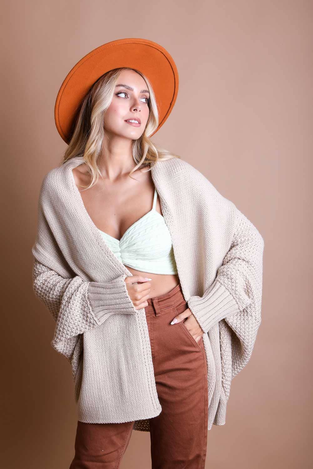 Woman wearing a beige cardigan, light green top, brown pants, and orange hat against a beige background