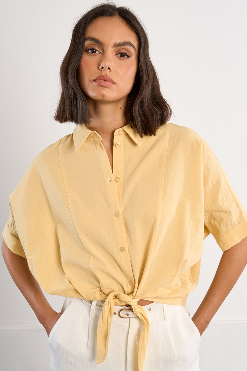Woman wearing a yellow button-up shirt with a tied front, standing against a plain background.