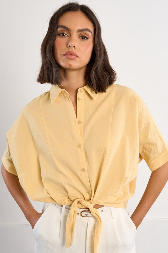 Woman wearing a yellow button-up shirt with a tied front, standing against a plain background.