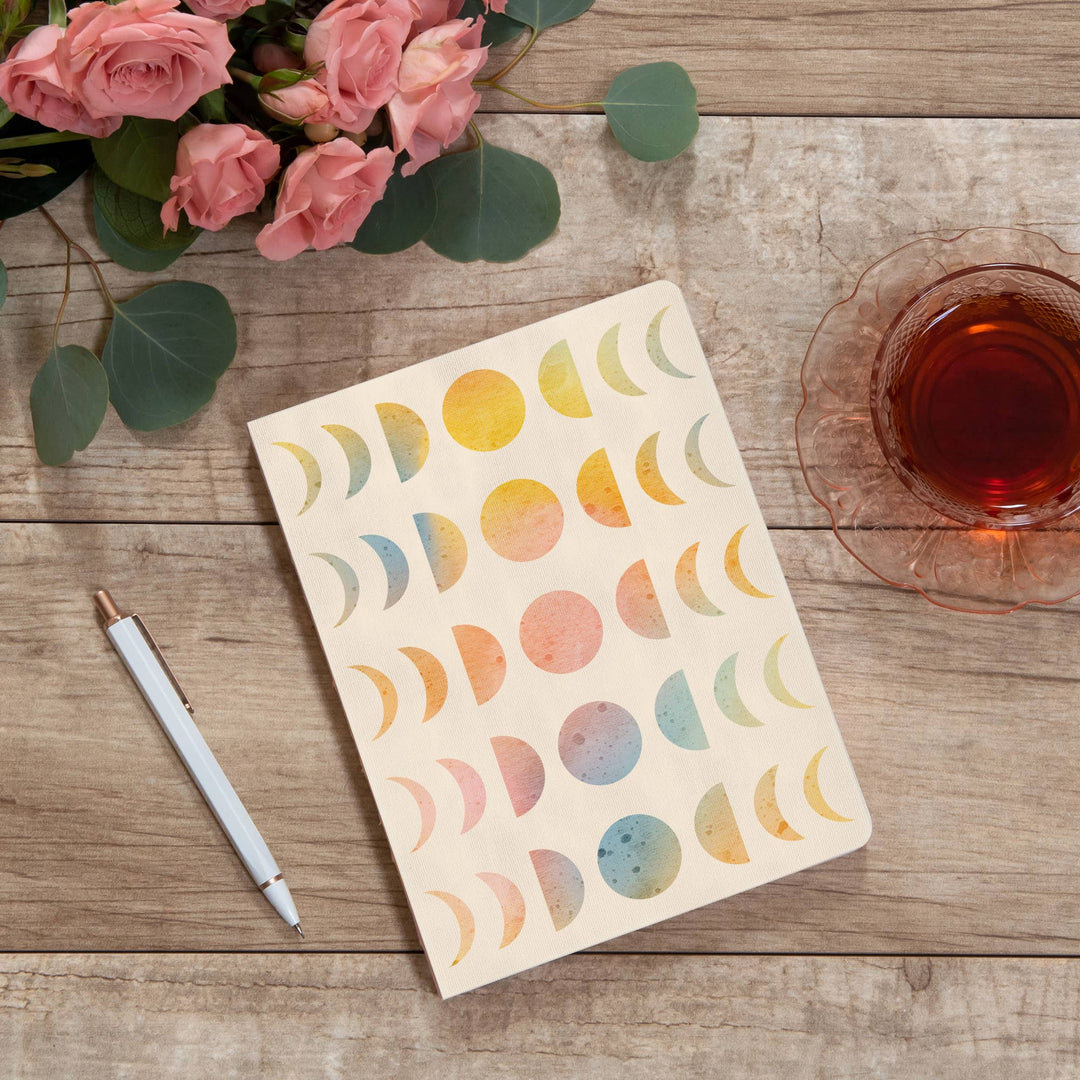 Notebook with colorful moon phases on a wooden surface with flowers and tea.