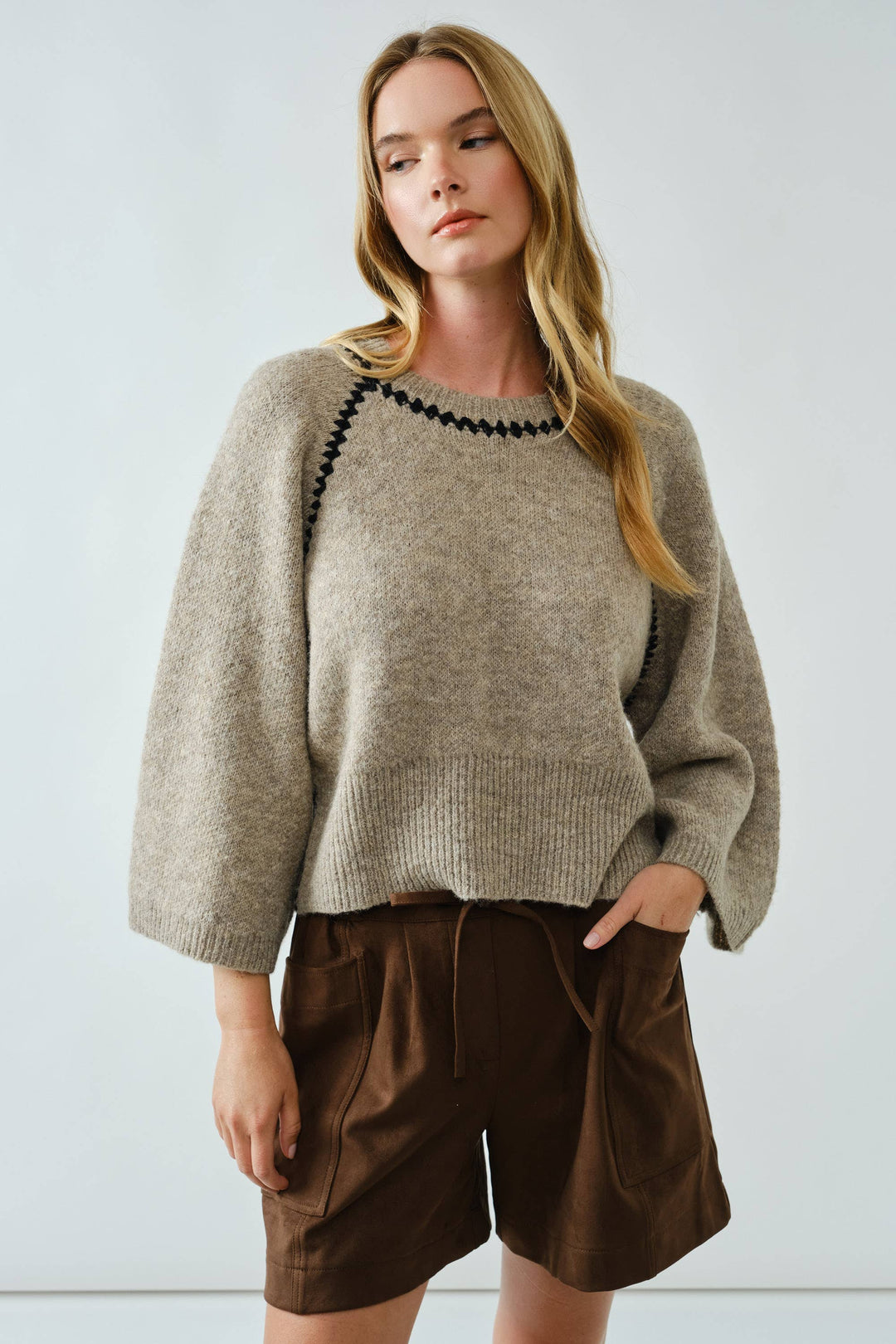 Woman wearing a beige sweater with black trim and brown shorts against a light background