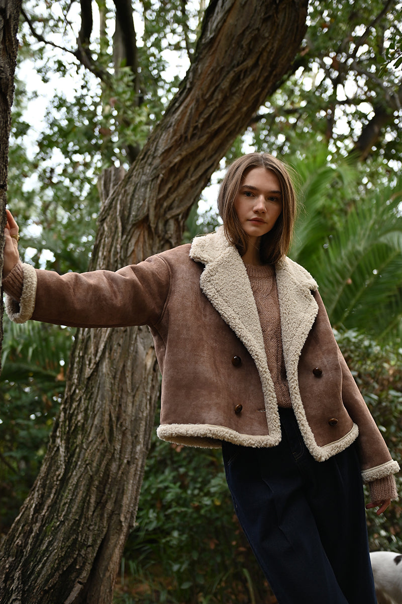 Person wearing a brown shearling jacket standing next to a tree in a forest setting