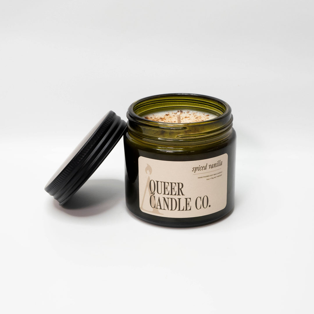 Candle jar with lid open, displaying candle inside, on a white background. Brand 'Queer Candle Co.' visible.