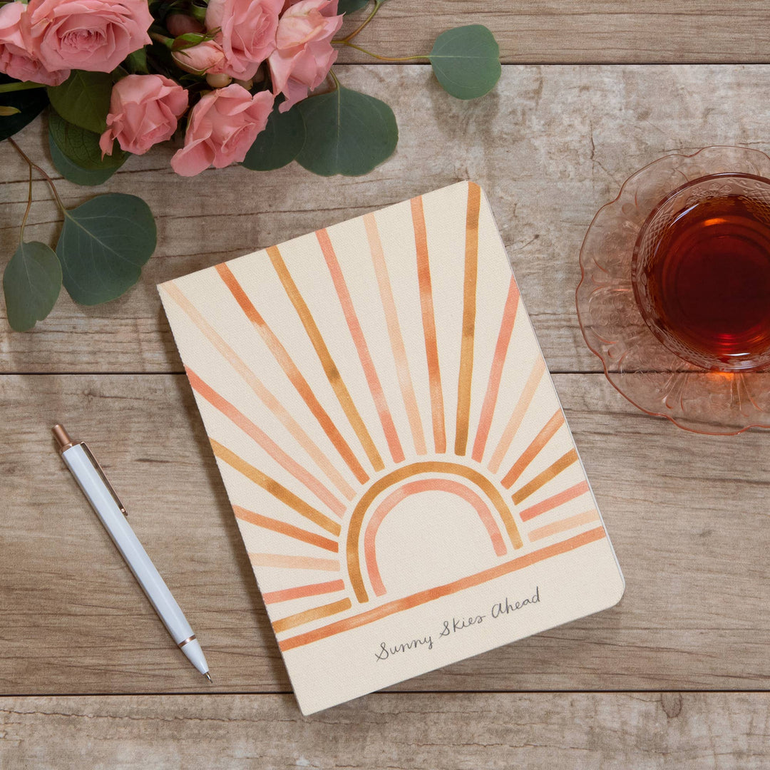 Notebook with sun design and 'Sunny Skies Ahead' text on a wooden surface with flowers and tea.